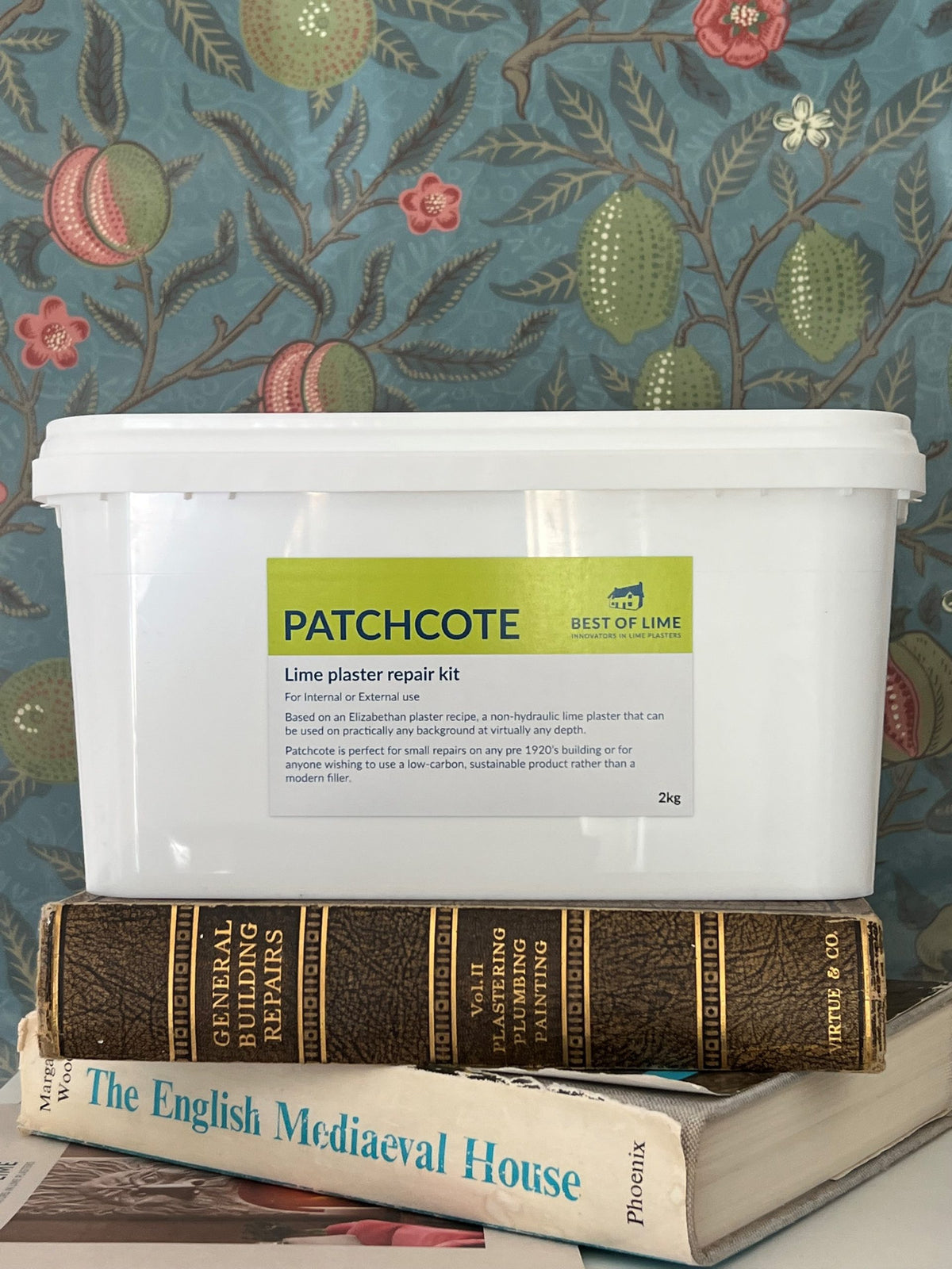 A white plastic container labeled 'PATCHCOTE' Plaster Repair Kit, placed on top of a book with the title 'The English Mediaeval House'. The container is set against a wallpaper with a floral pattern.